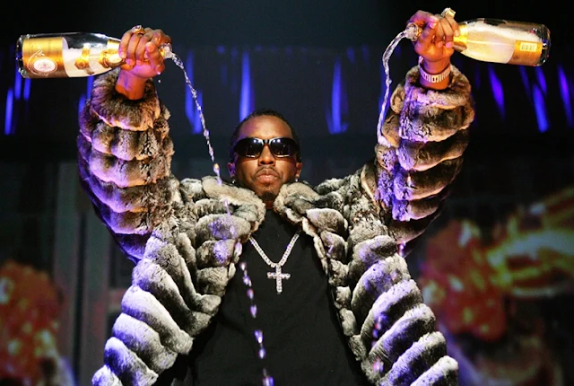 Diddy wearing an oversized fur coat with arms raised on stage, serving iconic fashion and hip-hop mogul energy.